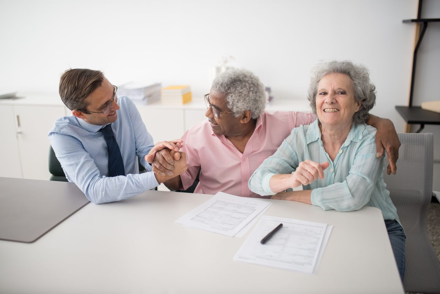 Consultor financeiro apertando a mão de um casal de clientes sorridentes durante uma reunião, com documentos e caneta sobre a mesa, representando a relação de confiança entre o profissional e os clientes