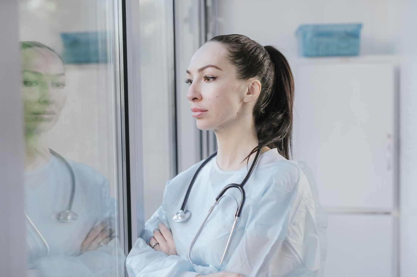 Médica de avental e estetoscópio, de braços cruzados, olhando pela janela de um corredor de clínica, com o próprio reflexo aparecendo no vidro ao lado dela