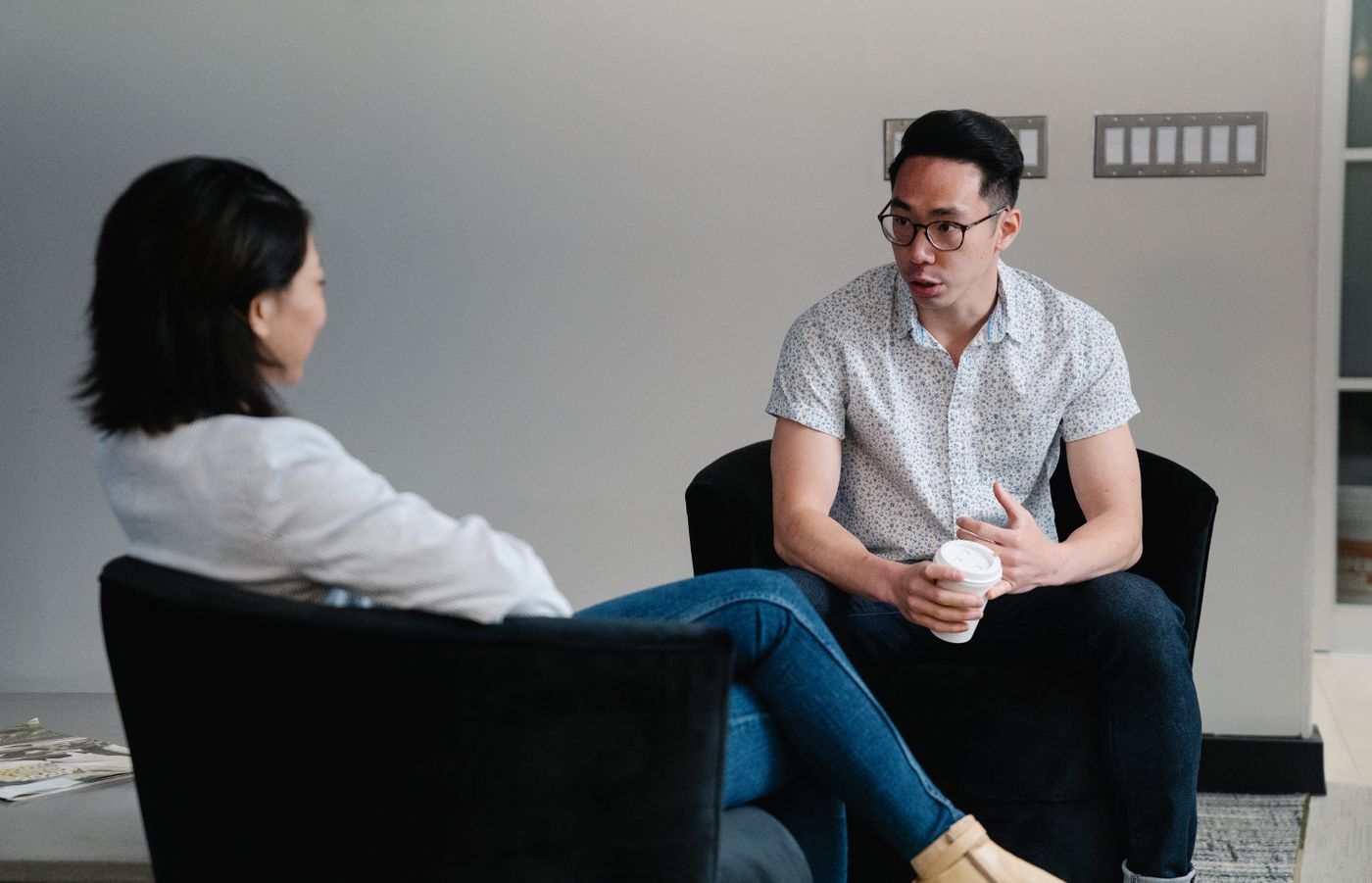 Duas pessoas sentadas em poltronas, uma delas falando com as mãos e segurando um copo de café enquanto a outra escuta, representando a conversa que conduz alguém até a sessão de aconselhamento