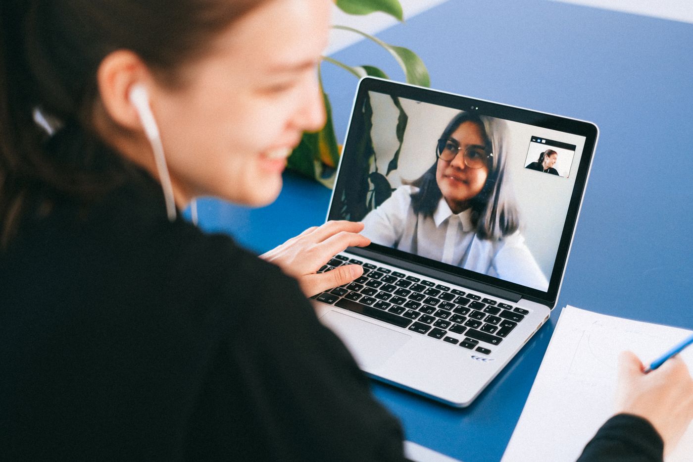 Mulher de costas, sorrindo, numa chamada de vídeo pelo notebook sobre uma mesa azul, com a cliente aparecendo na tela, representando o último encontro da consultoria feito por chamada