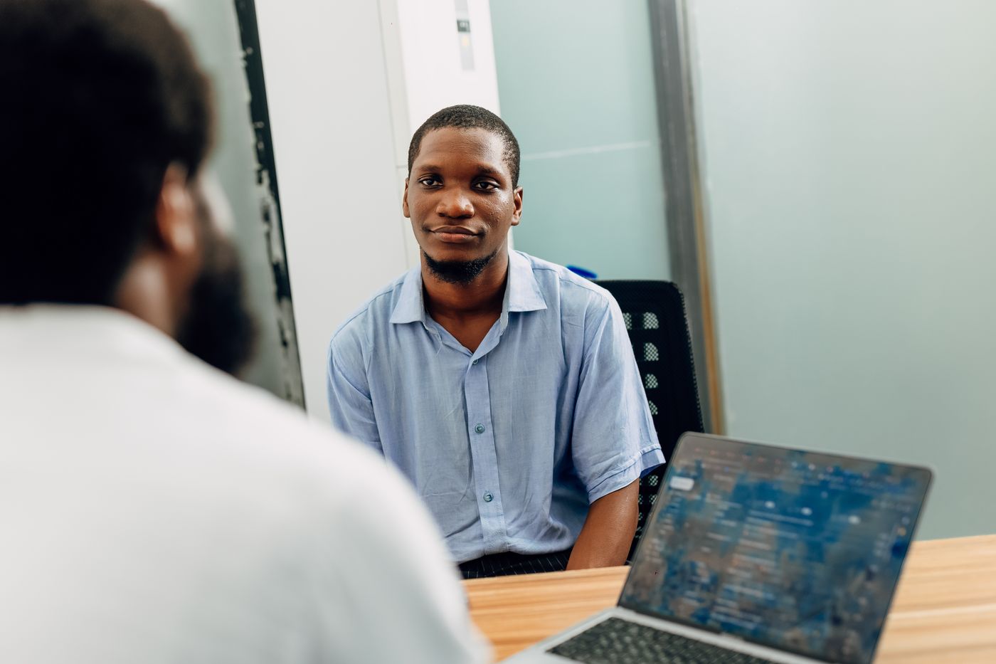 Dois homens sentados frente a frente numa mesa de escritório, um deles de costas em primeiro plano e o outro de camisa azul olhando diretamente para ele, com um notebook aberto ao lado, representando a conversa de cobrança entre o educador e o cliente