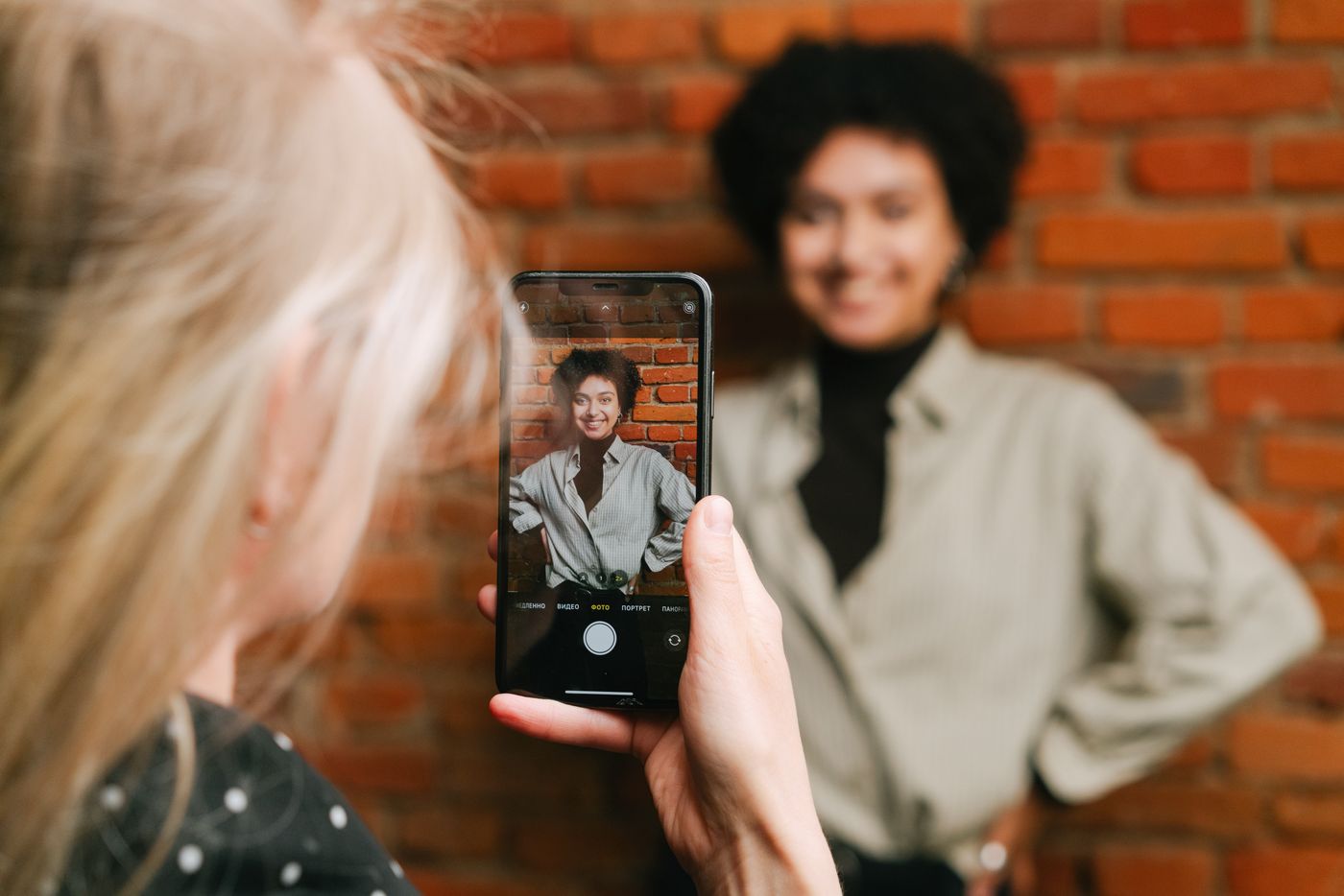 Mão segurando o celular que fotografa uma mulher sorrindo, de camisa clara e blusa de gola alta, diante de uma parede de tijolos, com o retrato dela aparecendo nítido na tela, representando a foto de perfil tirada com o próprio celular