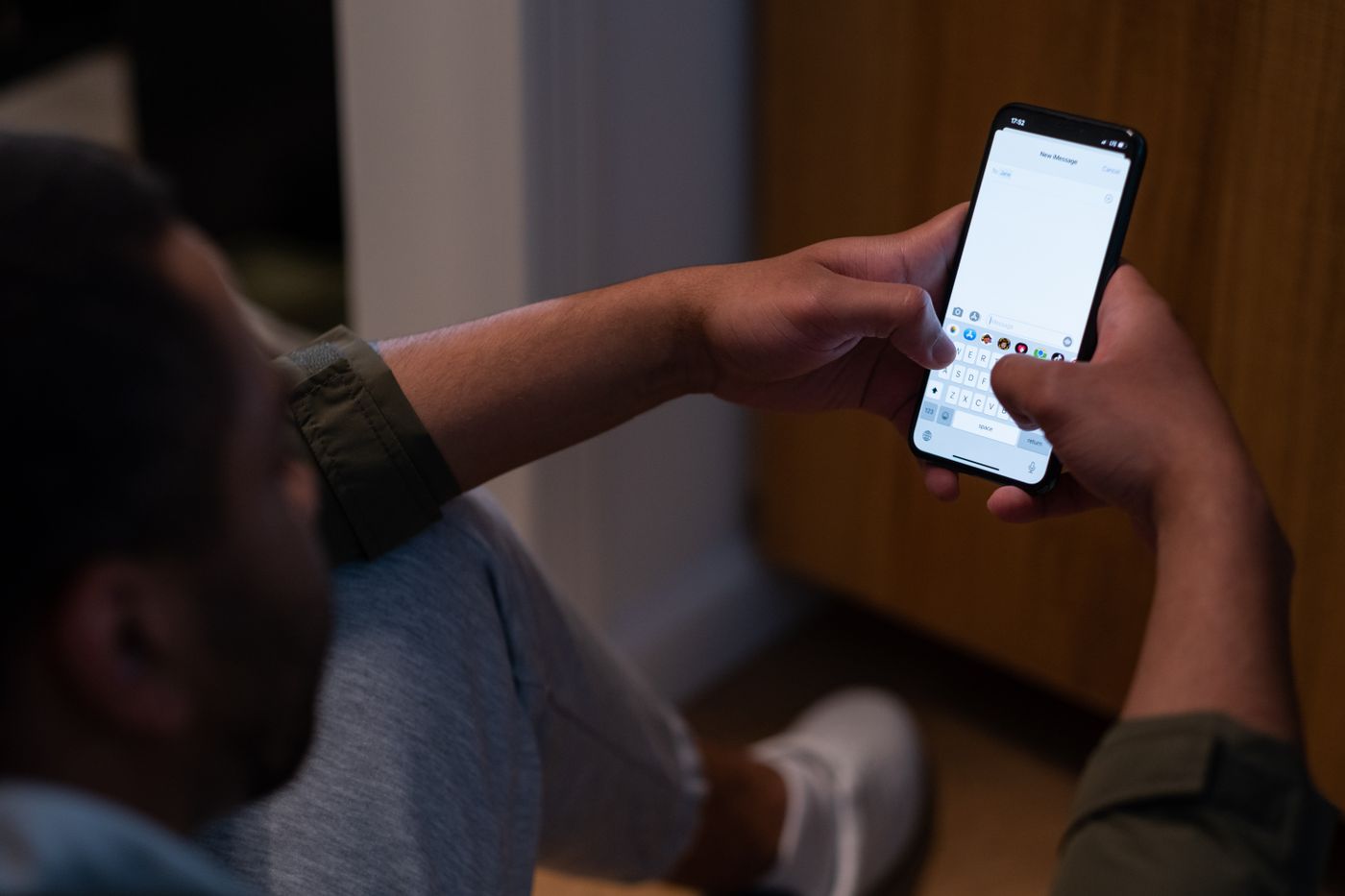 Vista por cima do ombro de um homem sentado num ambiente escuro, com as duas mãos no celular e o teclado aberto na tela de uma mensagem nova, representando o educador escrevendo para a própria rede de contatos