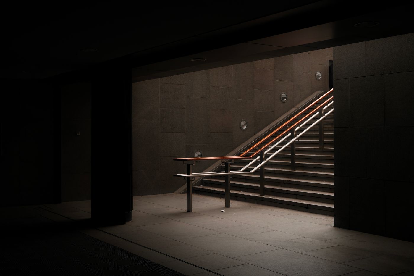 Escada de concreto subindo num corredor escuro, com o corrimão iluminado por uma linha de luz alaranjada e os degraus aparecendo um a um na penumbra, representando os três degraus da esteira de serviços