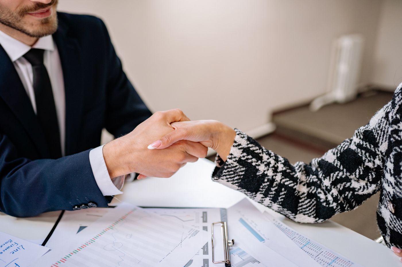 Duas pessoas de roupa social apertando as mãos sobre uma mesa coberta de relatórios financeiros impressos, com gráficos e planilhas espalhados, representando o acordo entre dois profissionais que atendem o mesmo cliente