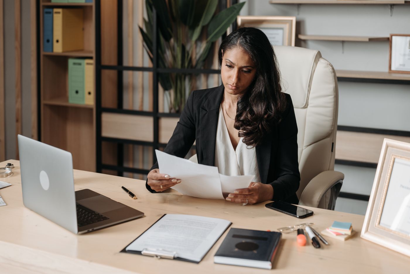 Profissional feminina em blazer revisando documentos na mesa com laptop, representando a decisão entre faculdade e formação específica para ser Educadora Financeira