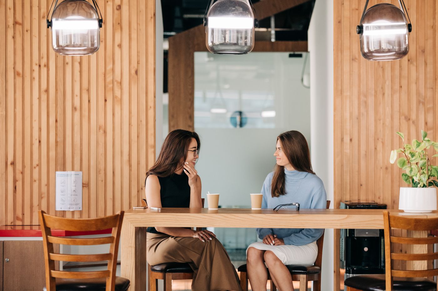 Duas mulheres sentadas lado a lado numa bancada de madeira clara, com copos de café à frente, conversando de perfil num espaço amplo com luminárias penduradas, representando a conversa em que o cliente conta o que mudou na vida dele