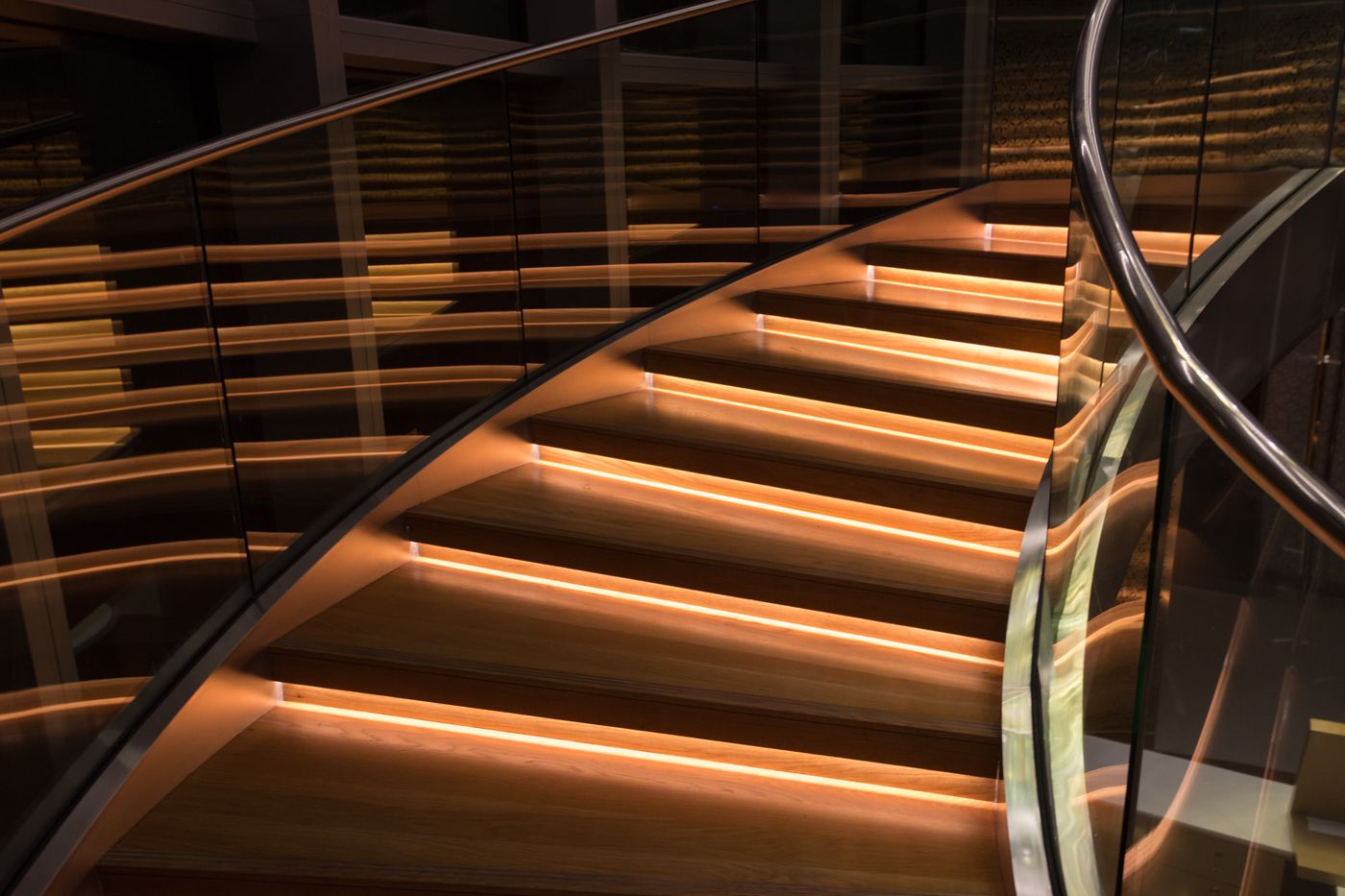 Escada curva de degraus de madeira iluminados por faixas de luz laranja, com corrimão de vidro, subindo em direção ao alto, representando o preço que sobe um degrau de cada vez