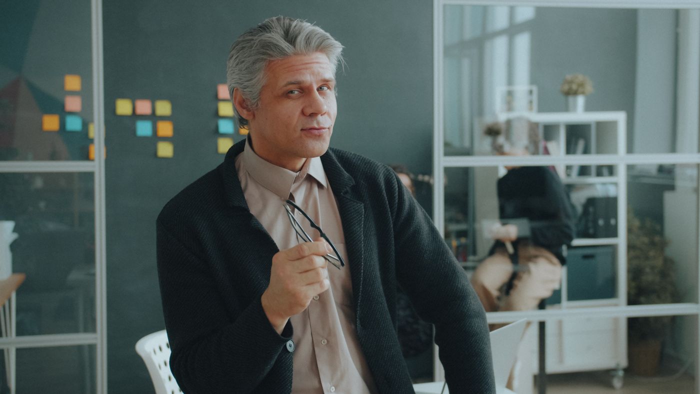 Homem grisalho de blazer escuro sentado num escritório aberto, segurando os óculos na mão e olhando para o lado com expressão tranquila, com uma parede de notas coloridas ao fundo, representando o profissional experiente que decide recomeçar
